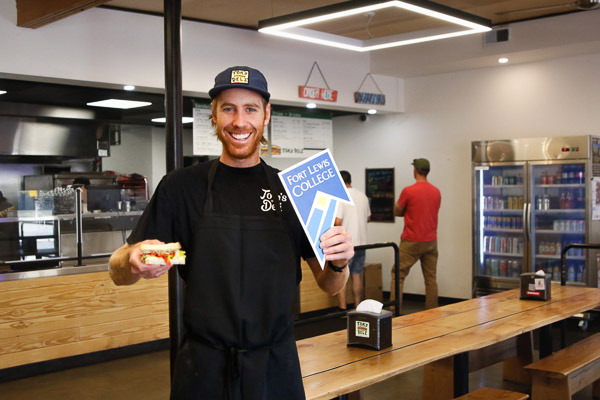 Tom holding a sandwich and a Fort Lewis College sign